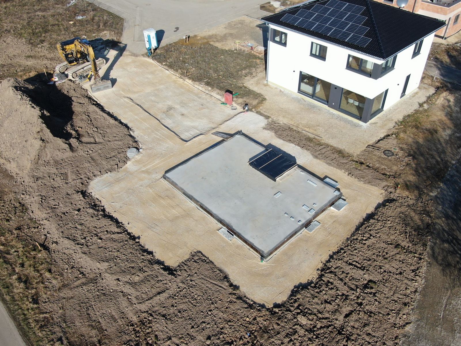 Luftbild einer fertigen Baugrube mit Bodenplatte neben einem Neubau